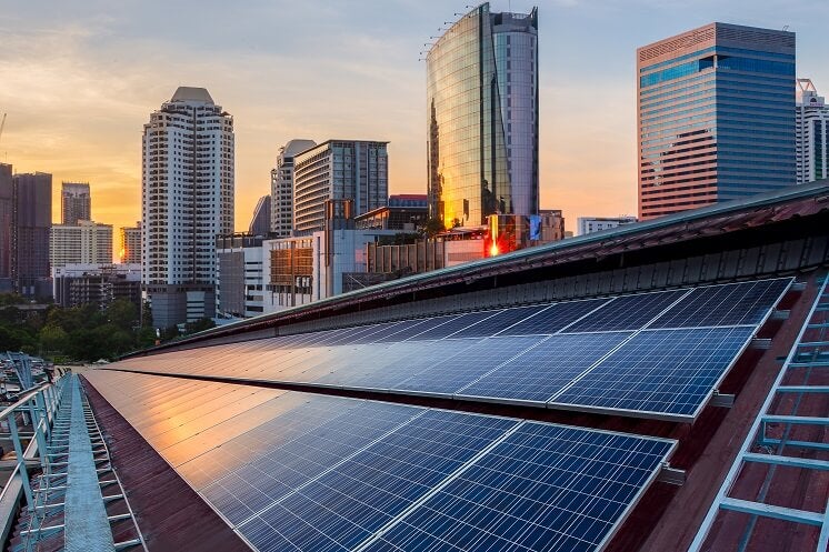 Photograph showcasing solar panels atop a brown building at sunset, with a cityscape of towering buildings visible in the soft evening light in the background, underscoring the integration of renewable energy within an urban setting.